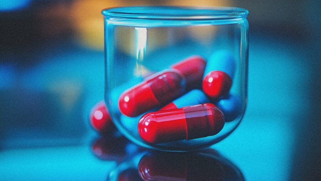 clean, modern close-up of red and blue capsules in a clear glass against a cool blue background with reflective surface